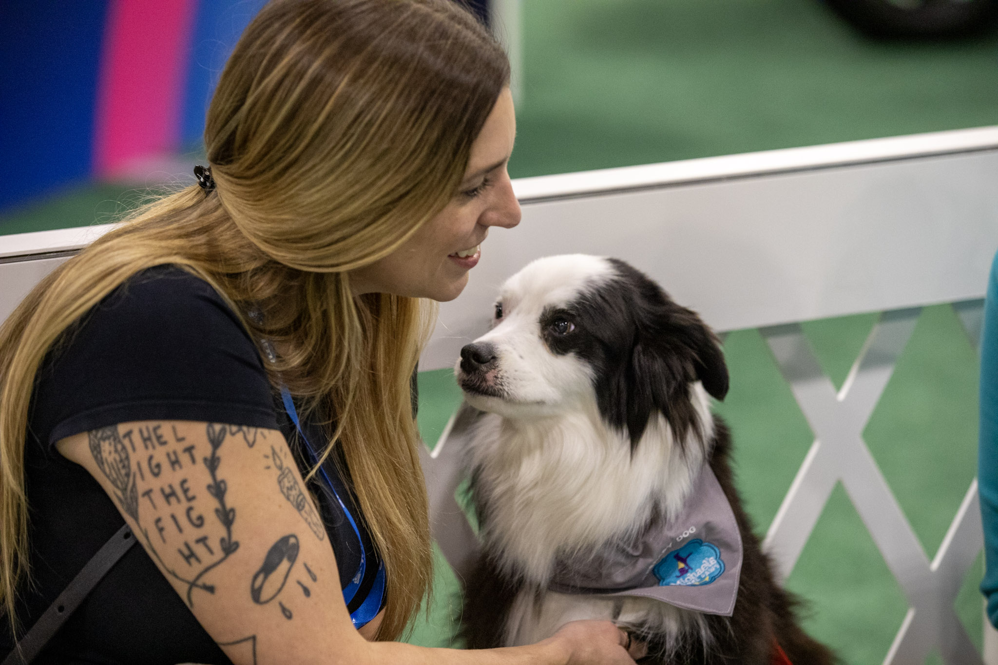 A woman with long blonde hair and arm tattoos smiles affectionately at a black and white dog wearing a grey bandana. They are positioned close together next to a white lattice fence with green flooring in the background.