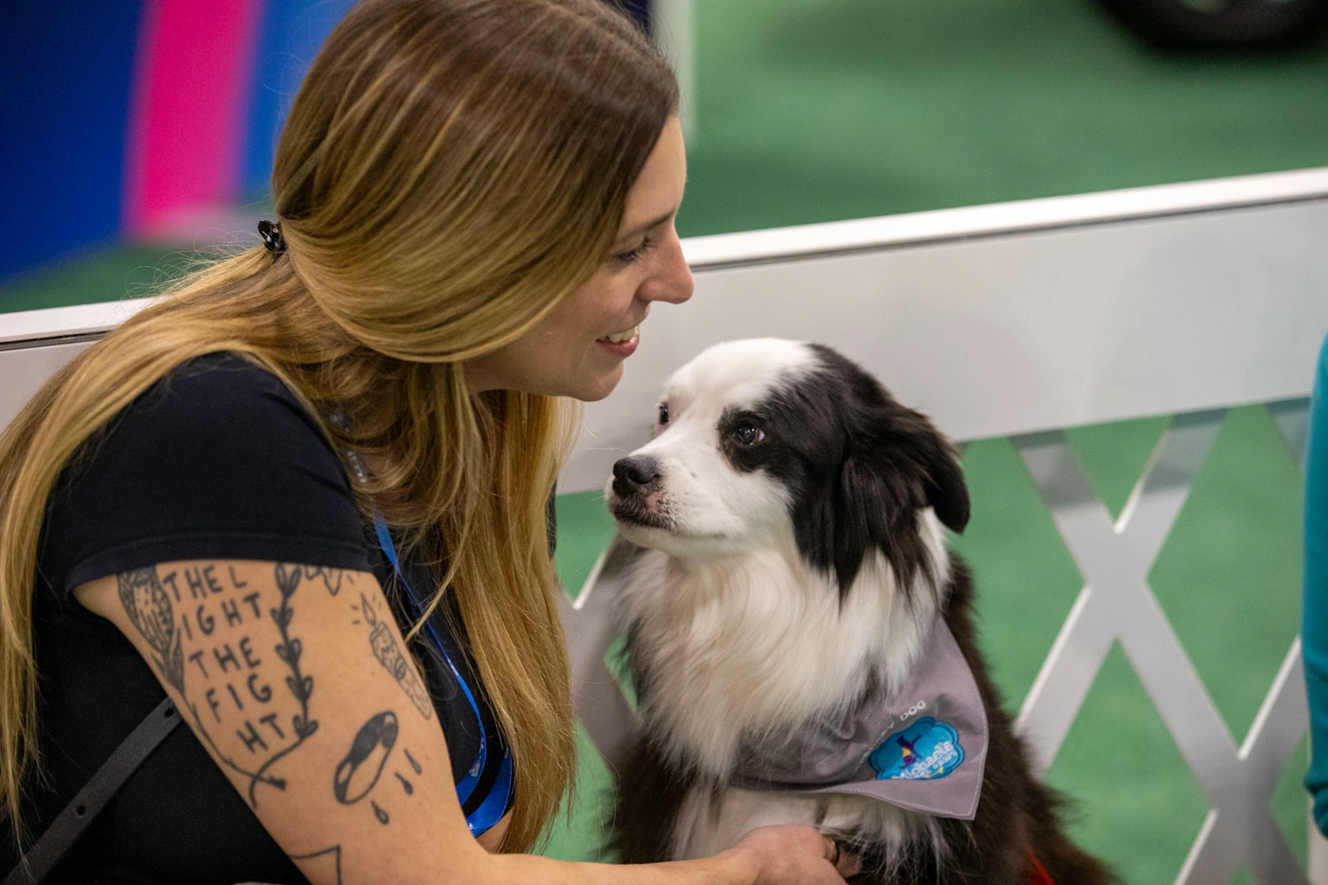 A woman with long blonde hair and arm tattoos smiles affectionately at a black and white dog wearing a grey bandana. They are positioned close together next to a white lattice fence with green flooring in the background.
