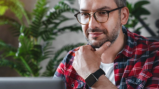 A bearded man in a plaid shirt and glasses touches his chin thoughtfully while looking at a laptop. Green plants are in the background.