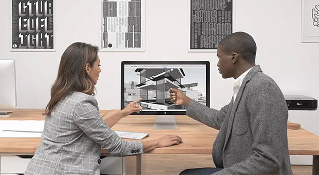 A man and a woman collaborate on an architectural 3D model using Trimble SketchUp software on a desktop monitor in a modern office.