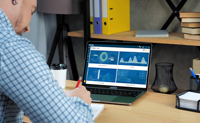 A man works at a desk, writing in a notebook while using a laptop displaying an e-Builder (a Trimble company) project management dashboard.