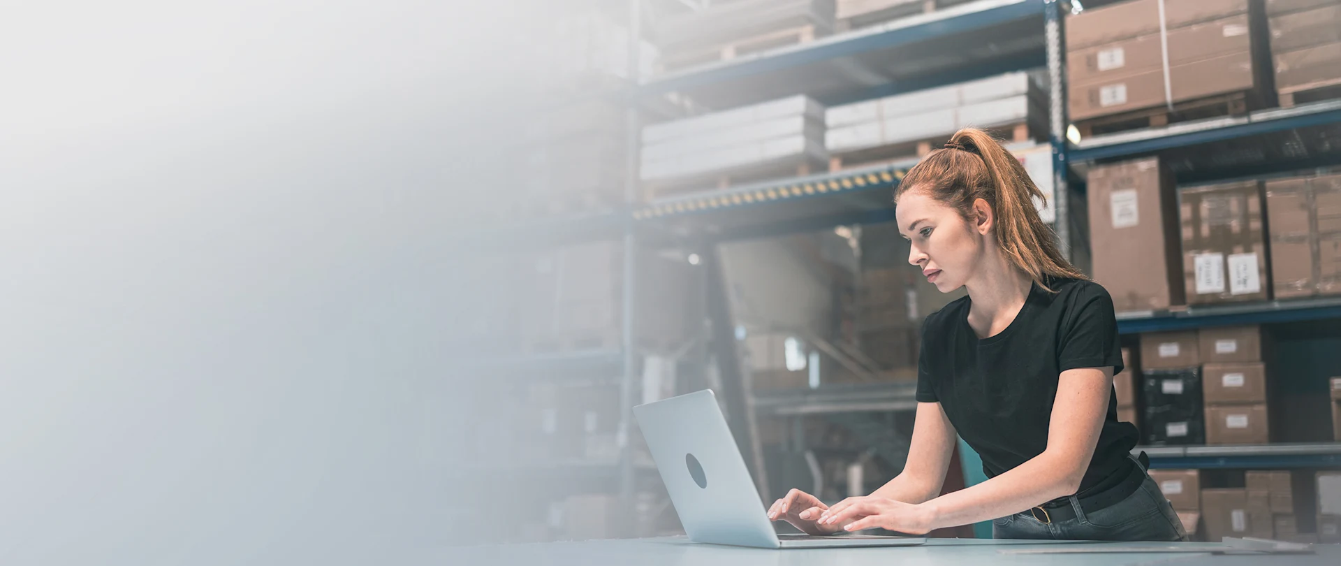 A woman with red hair in a ponytail works on a laptop at a table in a warehouse filled with shelves of cardboard boxes.