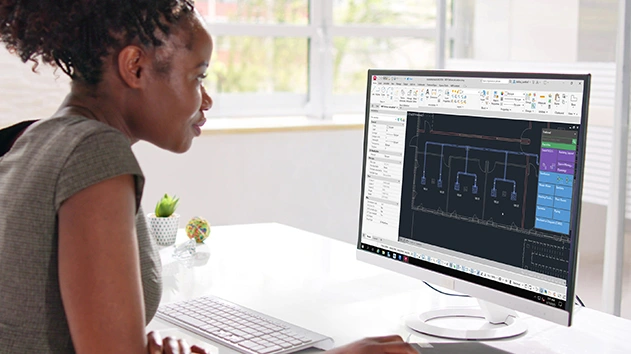 A woman in a bright office uses Stabicad for AutoCAD on a large monitor to design a mechanical building layout with blue ventilation or piping systems.