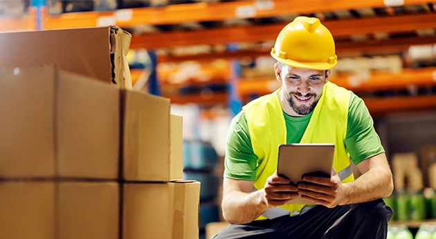 A warehouse worker in a yellow hard hat and safety vest using a tablet while crouched near stacks of cardboard boxes.