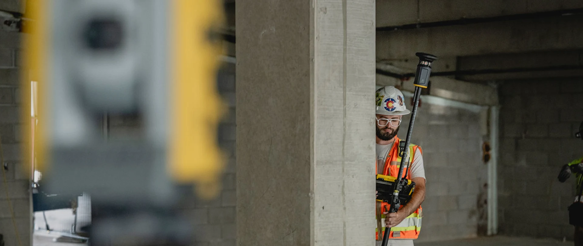 A building construction professional in a hard hat and safety vest operates a surveying rod and tablet in an unfinished building, with a total station blurred in the foreground.