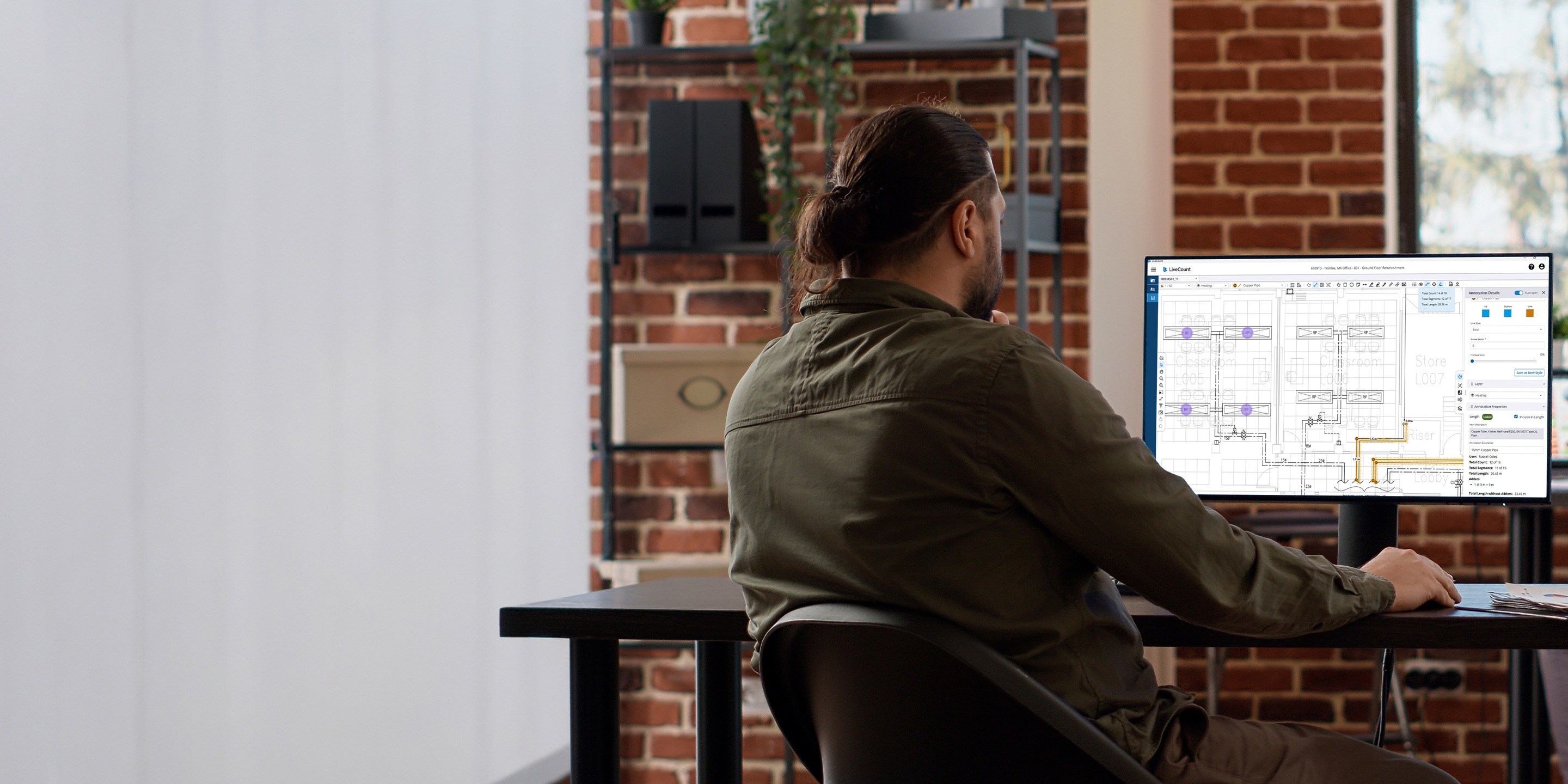 A man with long dark hair tied back sits at a desk, facing away. He is looking at a screen monitor displaying LiveCount software.