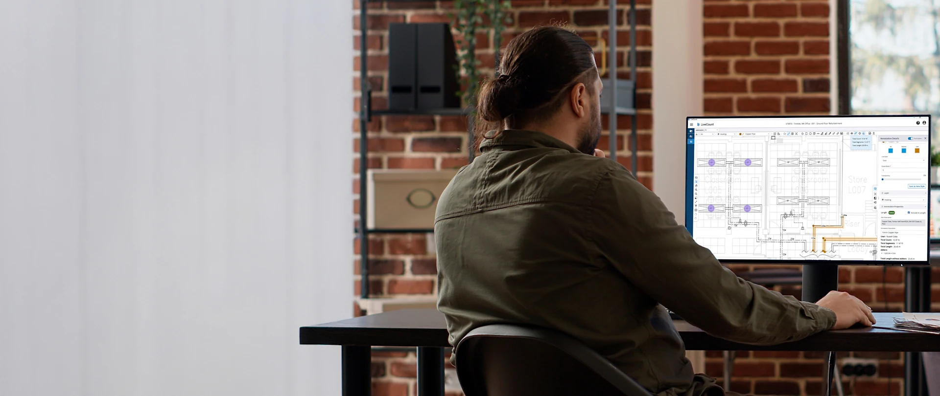 A man with long dark hair tied back sits at a desk, facing away. He is looking at a screen monitor displaying LiveCount software.