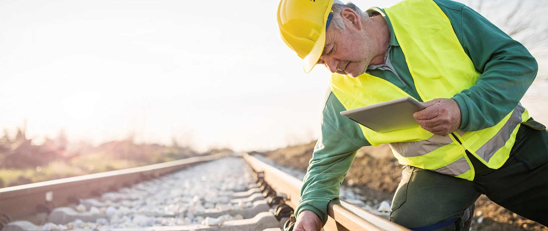 A worker in a yellow hard hat and vest inspects a train track while holding a tablet.