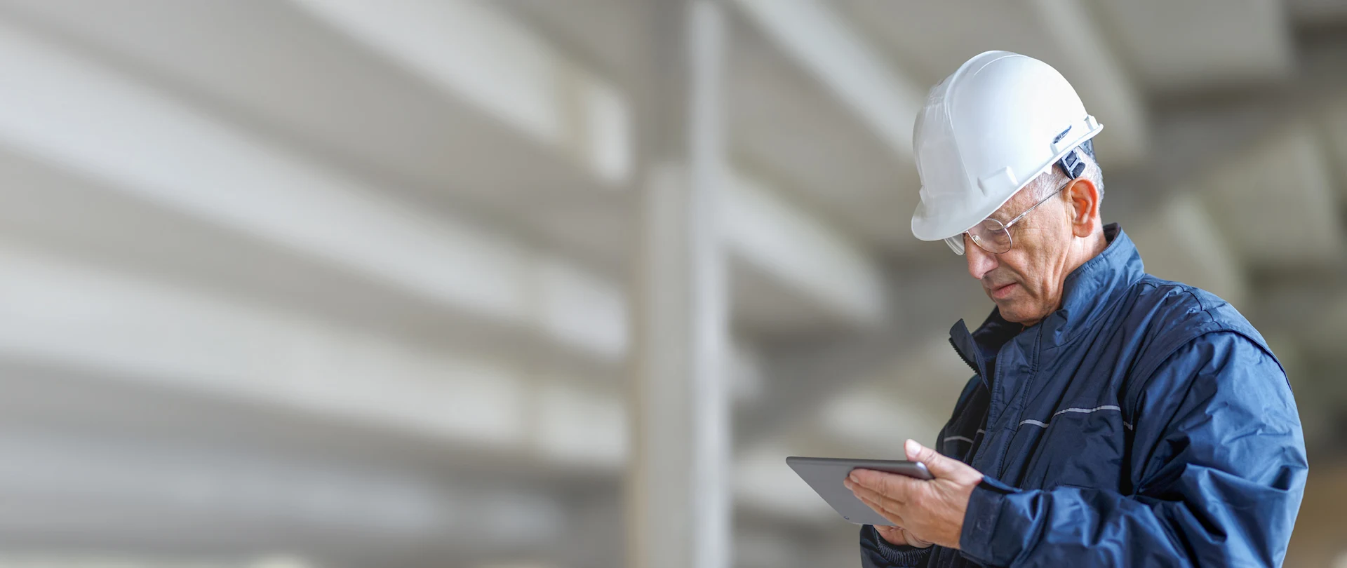 A senior construction professional in a white hard hat and blue jacket uses a tablet at a construction site.
