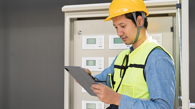 A technician in a yellow hard hat and safety vest uses a tablet to inspect an industrial control panel with digital displays.