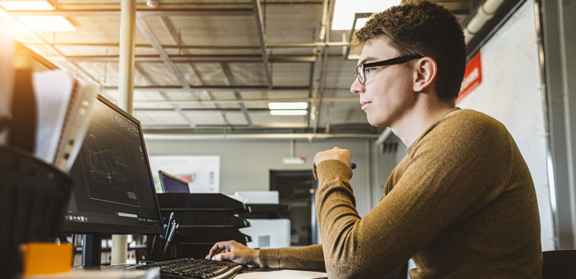 Man with yellow sweater at desk looking at computer showing graphs