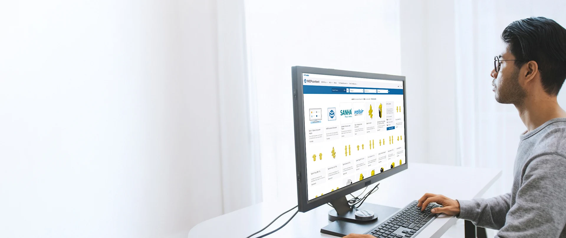 A person sits at a desk, focused on a computer monitor displaying a MEPcontent website with BIM objects.
