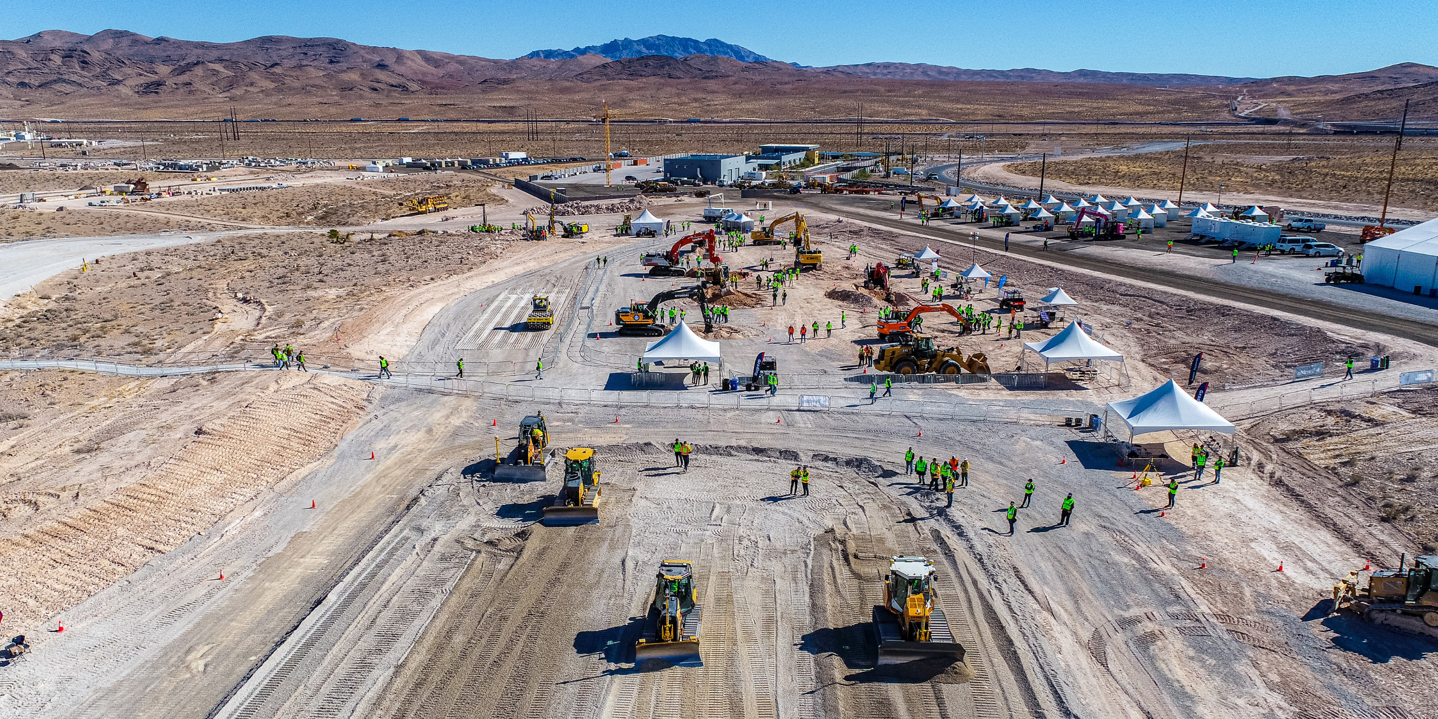Aerial view of an outdoor construction expo in a desert, featuring heavy machinery like bulldozers, excavators, white tents, and event attendees.