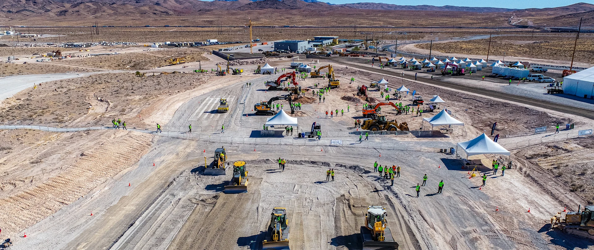 Aerial view of an outdoor construction expo in a desert, featuring heavy machinery like bulldozers, excavators, white tents, and event attendees.