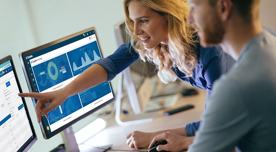 Two colleagues review a Trimble Supplier Xchange digital dashboard on a computer monitor.