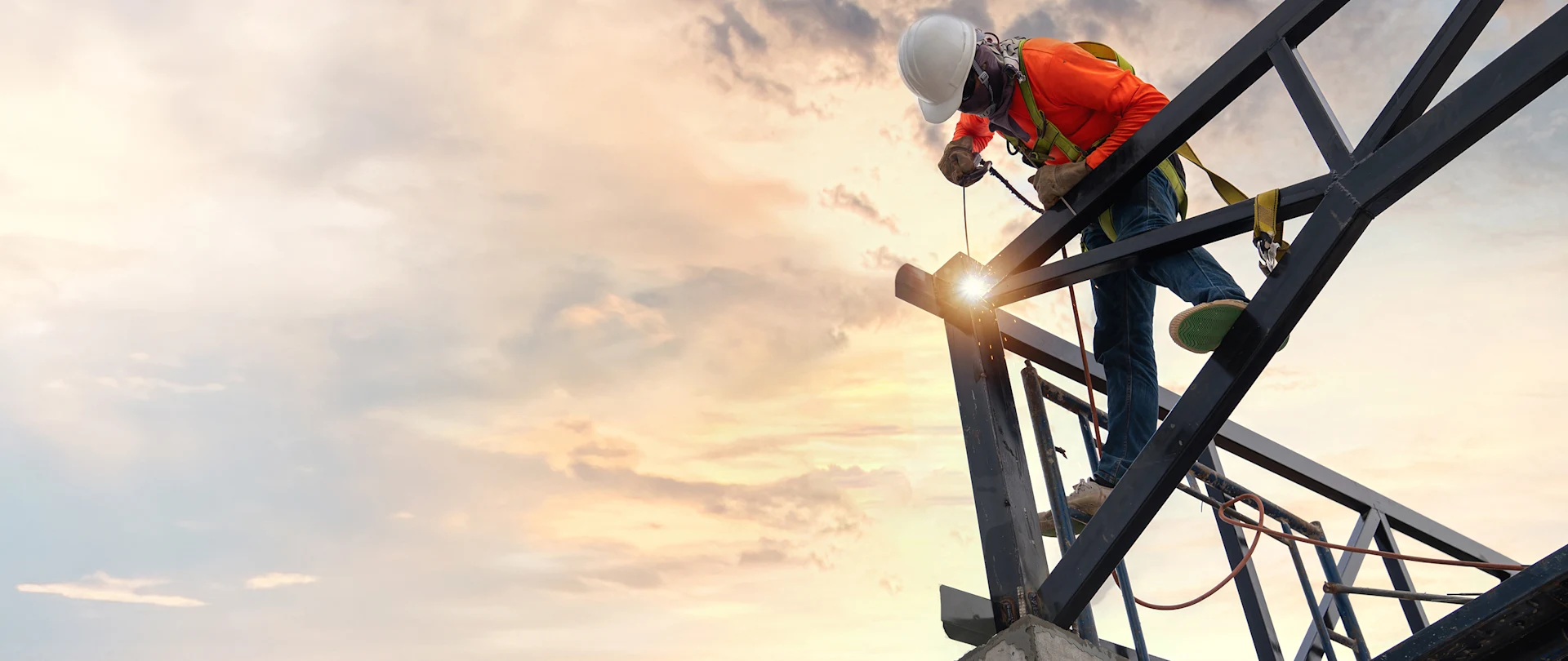 A construction worker in safety gear welds a steel structure against a sunset sky, with bright sparks flying from the welding tool.