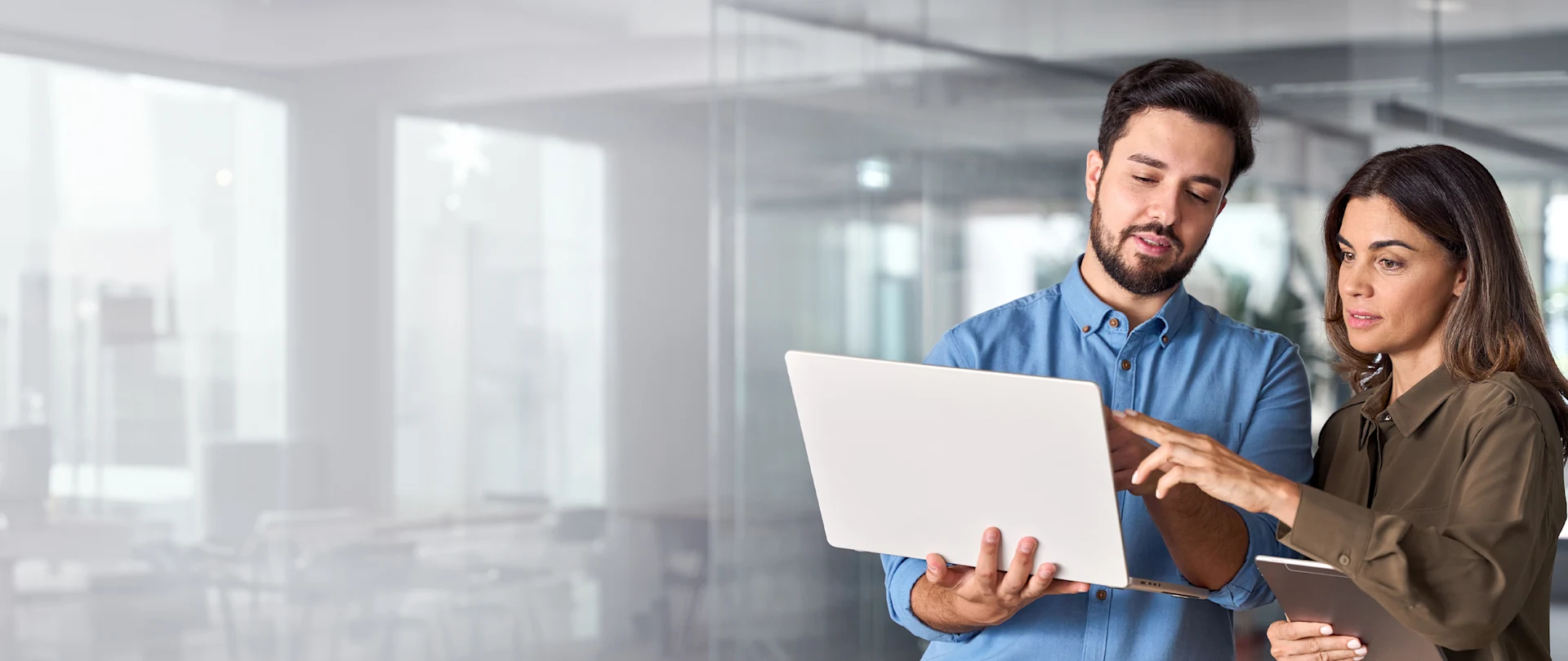 In this image, a man in a blue shirt and a woman in a brown shirt collaborate while looking at a laptop and digital tablet in a modern office setting.