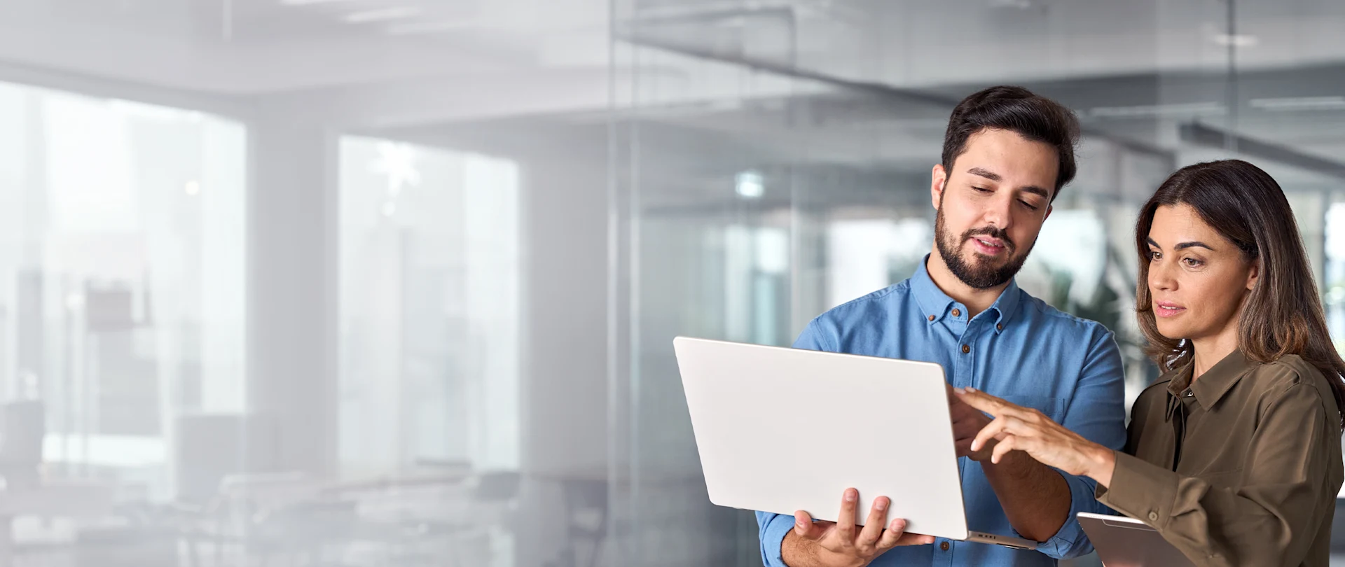 In this image, a man in a blue shirt and a woman in a brown shirt collaborate while looking at a laptop and digital tablet in a modern office setting.