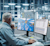 A man uses Trimble Tekla Structures software on dual monitors to design a steel model in a factory workshop environment.