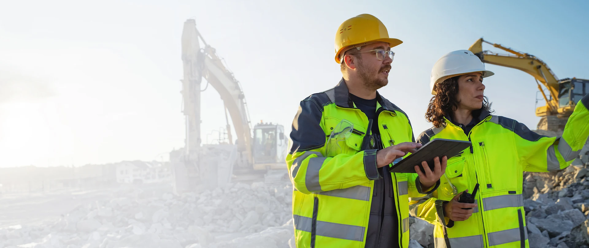 Two construction workers in safety gear use a tablet at a site with excavators, showcasing Trimble’s connected construction technology.