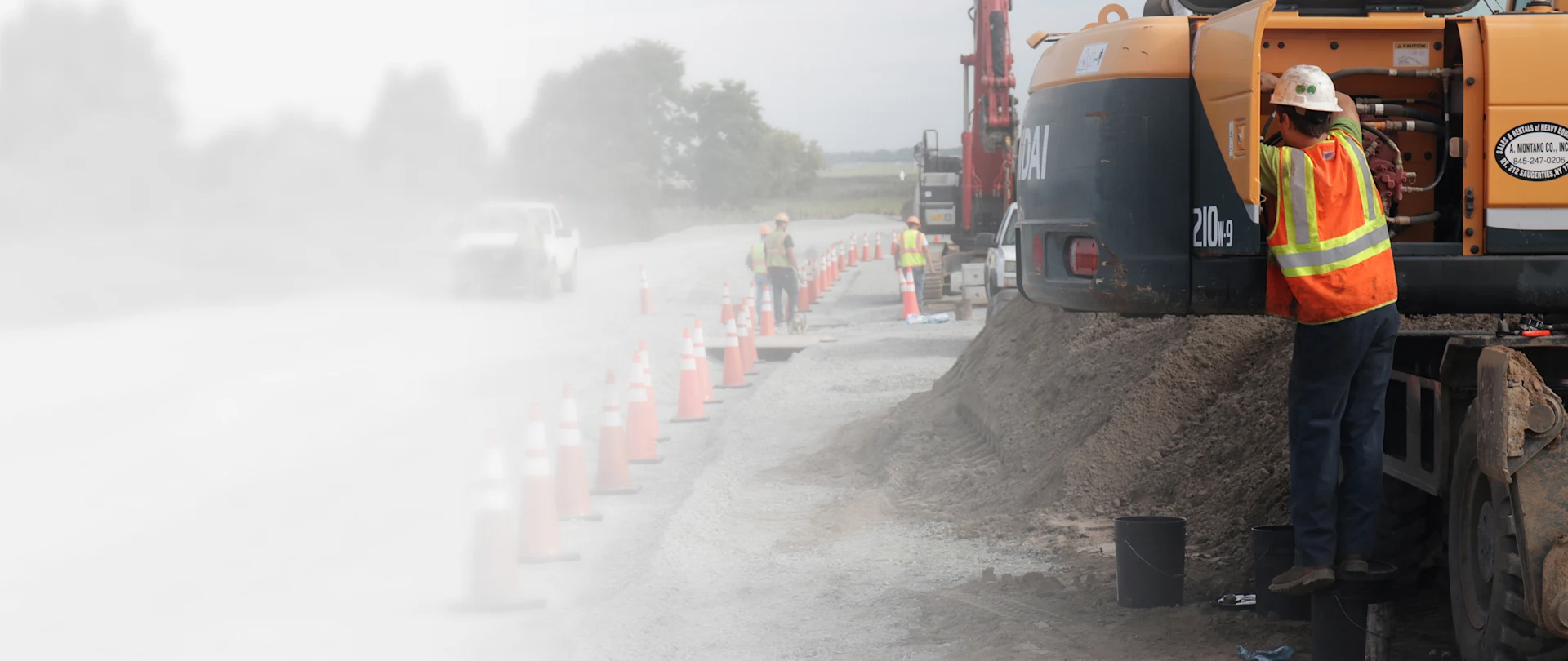 A worker in high-visibility safety gear services a Hyundai 210W-9 wheeled excavator on a dusty road construction site near Saugerties, New York.