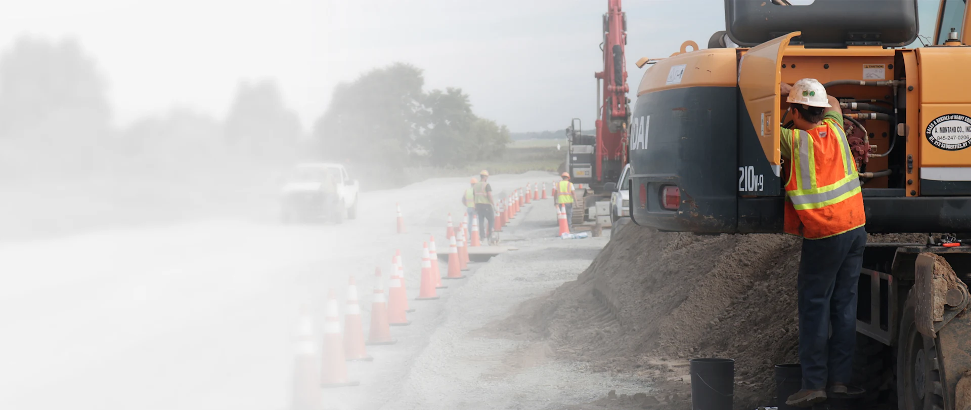 A worker in high-visibility safety gear services a Hyundai 210W-9 wheeled excavator on a dusty road construction site near Saugerties, New York.