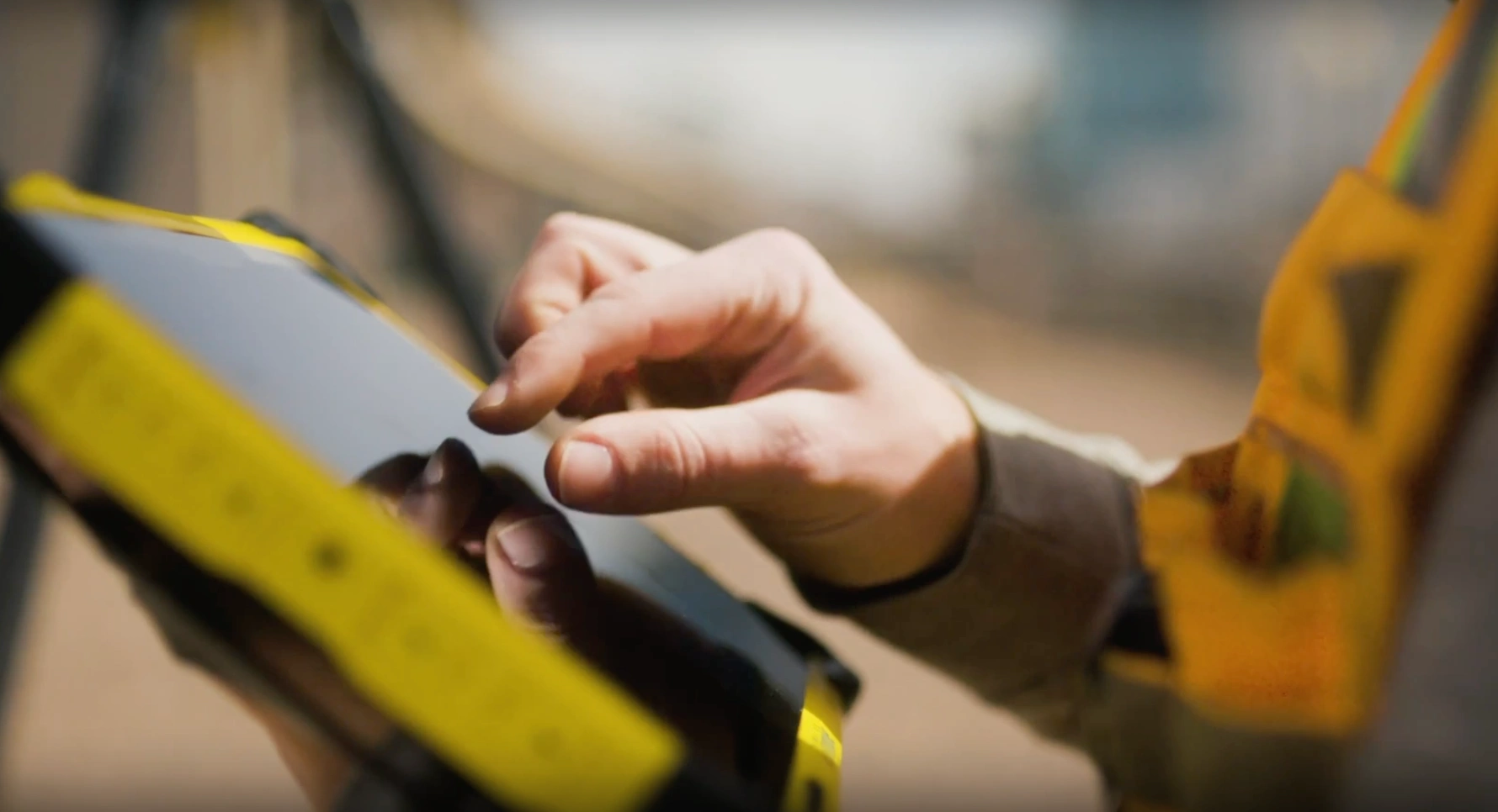 A close-up of hands using a yellow Trimble T110 rugged tablet on a field jobsite, with blurred survey equipment in the background.