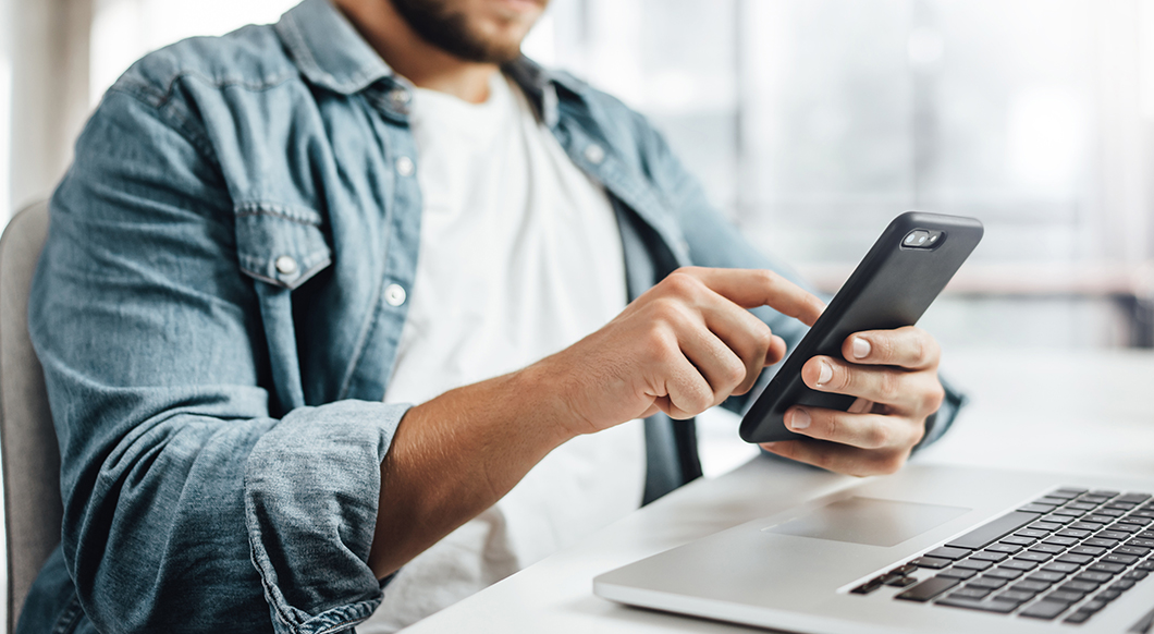 A man in a denim shirt and white t-shirt uses a smartphone while sitting at a laptop in a bright, modern office or workspace.