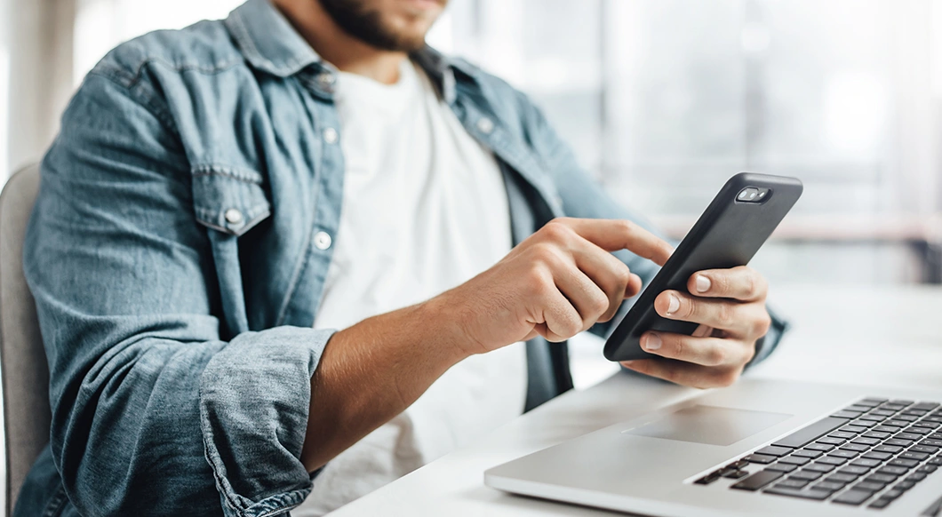 A man in a denim shirt and white t-shirt uses a smartphone while sitting at a laptop in a bright, modern office or workspace.