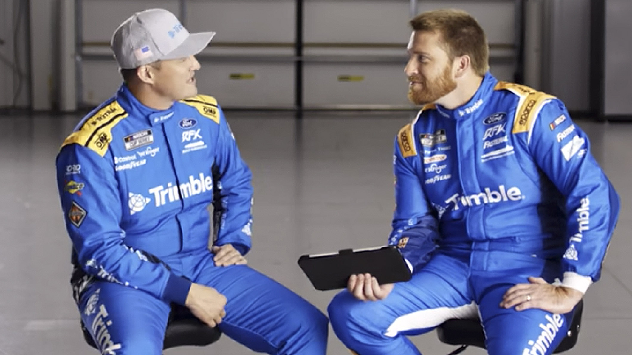 NASCAR drivers Ryan Preece and Chris Buescher in blue RFK Racing suits with Trimble sponsorship, discussing in a garage.