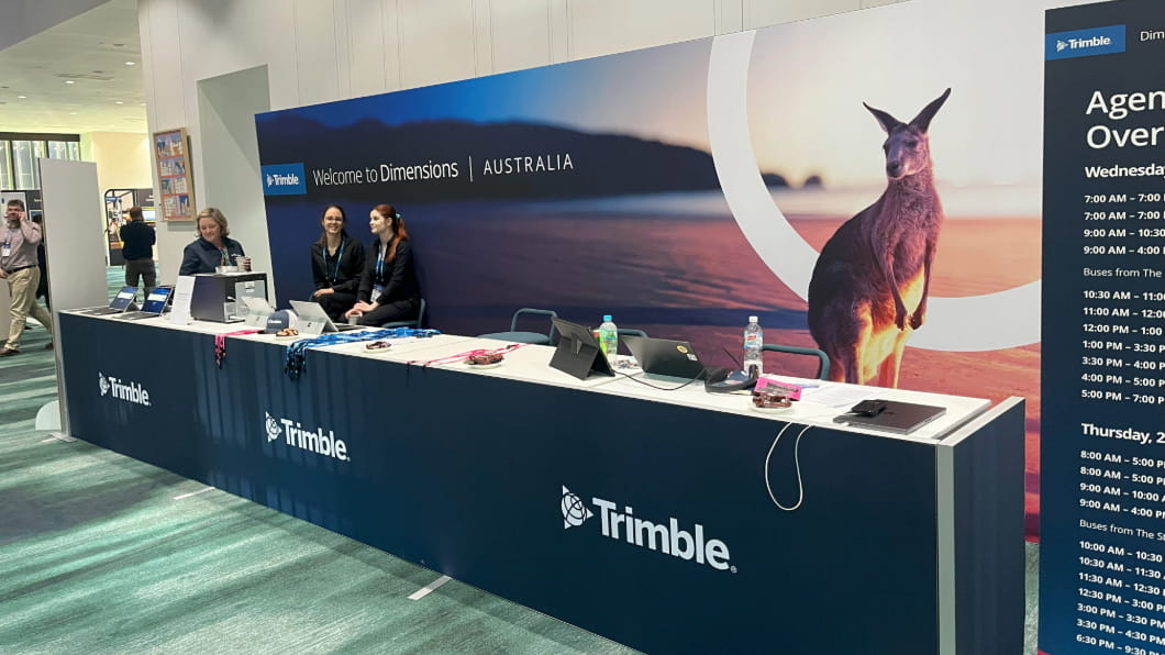 At the **Trimble Dimensions Australia** conference in **Brisbane**, staff assist attendees at a registration desk featuring **Trimble** branding and a kangaroo backdrop.