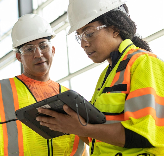 Two construction workers in hard hats and safety vests review data on a rugged Trimble tablet at a job site.