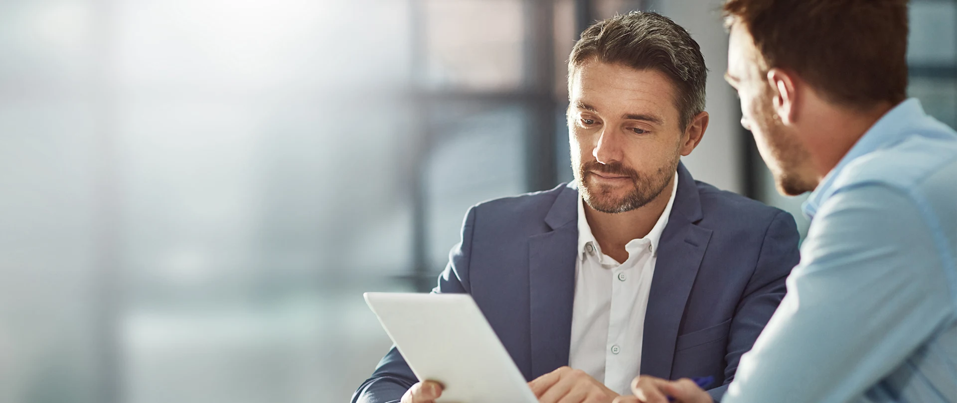 Two men in business attire review information together on a white tablet in a bright, modern office setting.