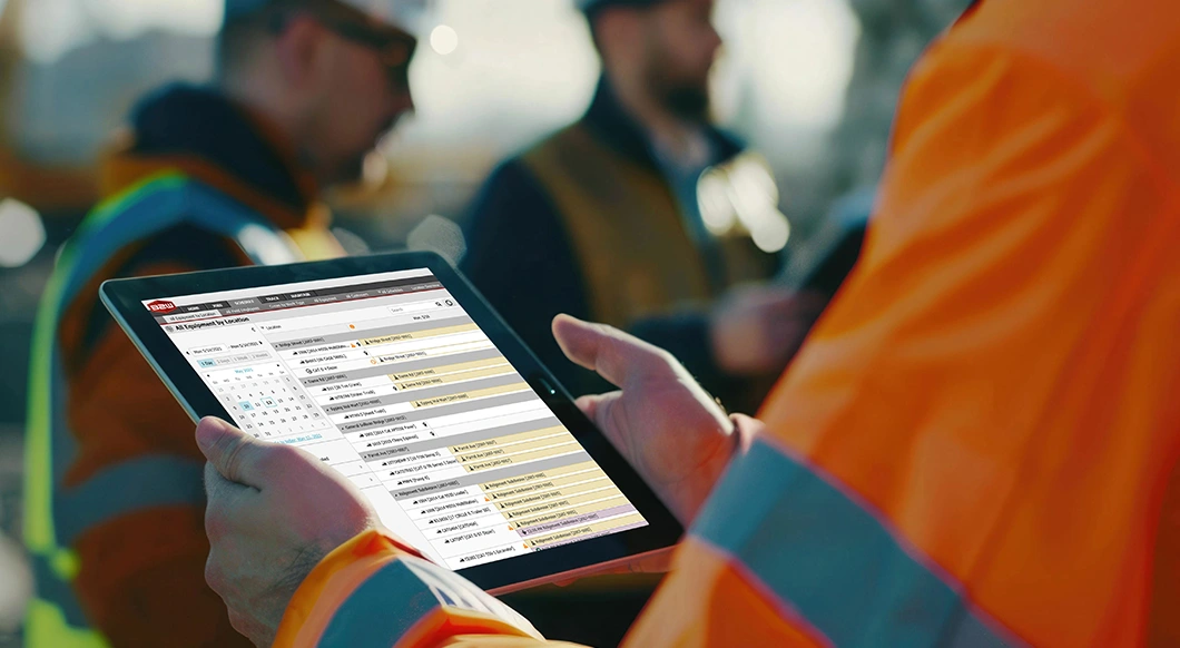 A person in a safety vest holds a tablet displaying Trimble's B2W Schedule software for construction equipment and resource management.