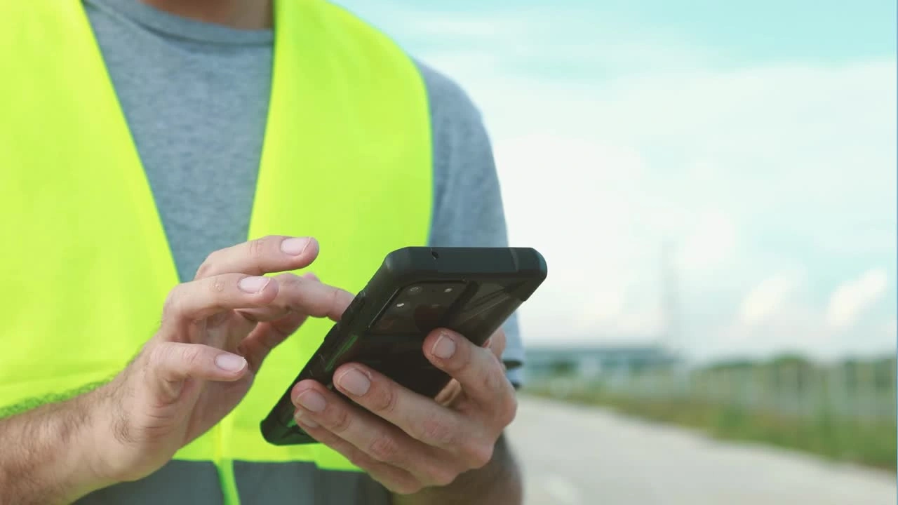 A construction worker in a high-visibility vest uses a smartphone to access Trimble field software on a roadside job site.
