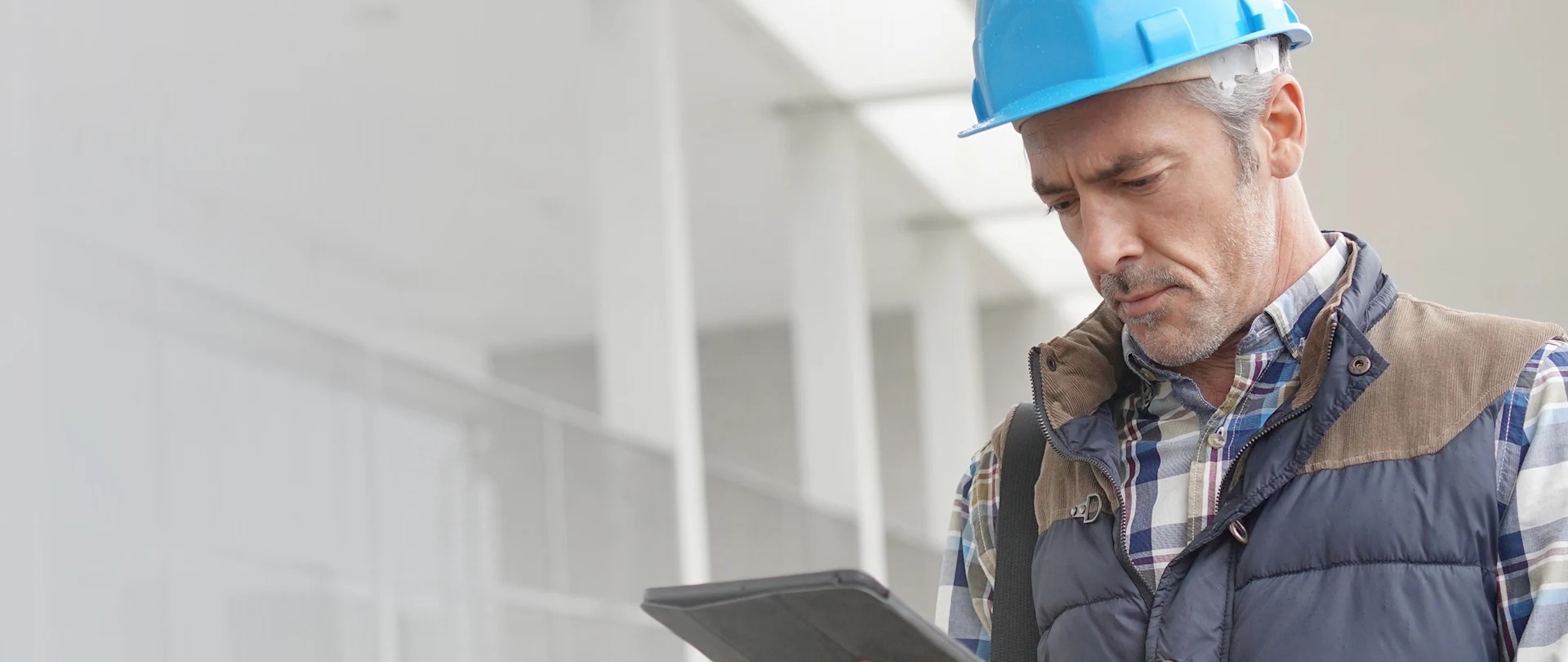 A worker in a blue hard hat and puffer vest uses a Trimble rugged tablet at a construction site.