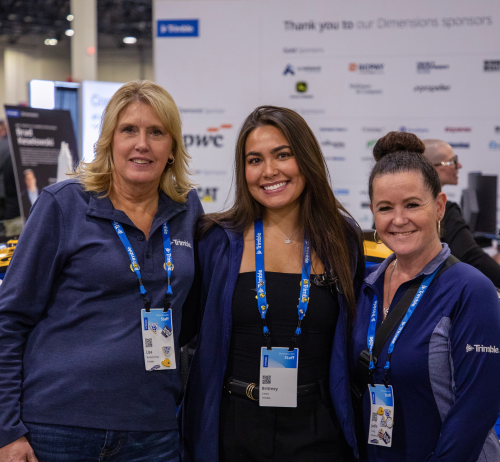 Three Dimensions employees in Trimble gear smile for the camera in front of the sponsor logo wall in the Expo hall.