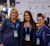 Three Dimensions employees in Trimble gear smile for the camera in front of the sponsor logo wall in the Expo hall.