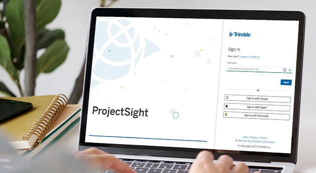 Hands typing on a laptop displaying the Trimble ProjectSight sign-in page, with a notebook and smartphone on a desk.