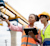 Two women in safety vests at a construction site; one points while holding a walkie-talkie and the other views a tablet near a large gantry crane.
