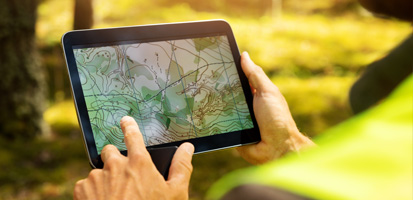Hands holding a tablet displaying a topographic map. The background is a sunny, blurred outdoor environment, suggesting a fieldwork context.