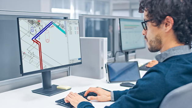 A man with curly hair and glasses works at a computer, looking at a 3D model of pipes on screen.