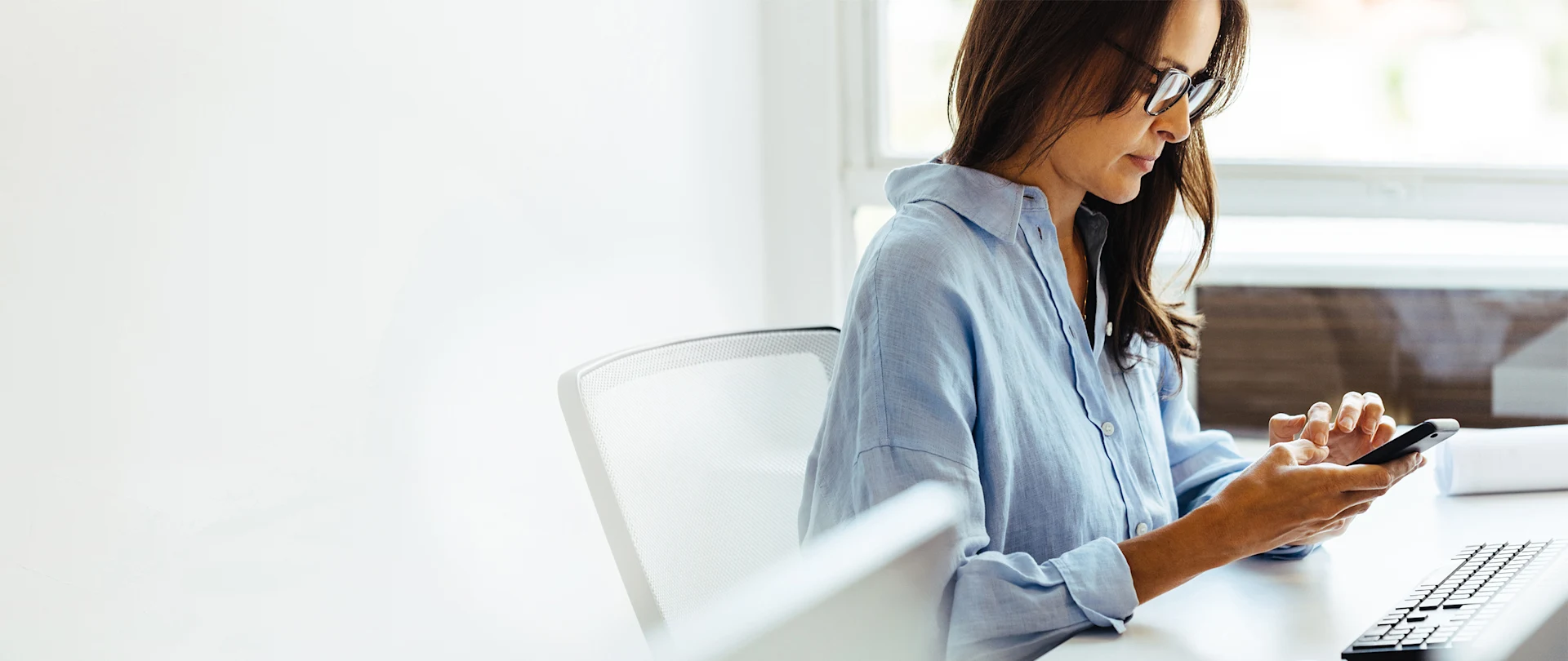 A professional woman wearing glasses and a blue shirt uses her smartphone at a white desk with a keyboard and mouse in a bright, modern office.