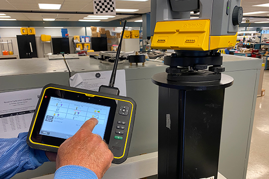 A person's hand operating a Trimble controller tablet, interacting with a Trimble surveying instrument in a laboratory or calibration setting.