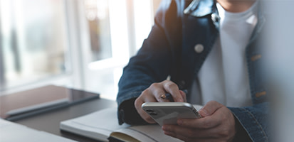 A person wearing a denim jacket uses a smartphone at a desk with an open notebook and a pen in a bright, modern professional workspace.