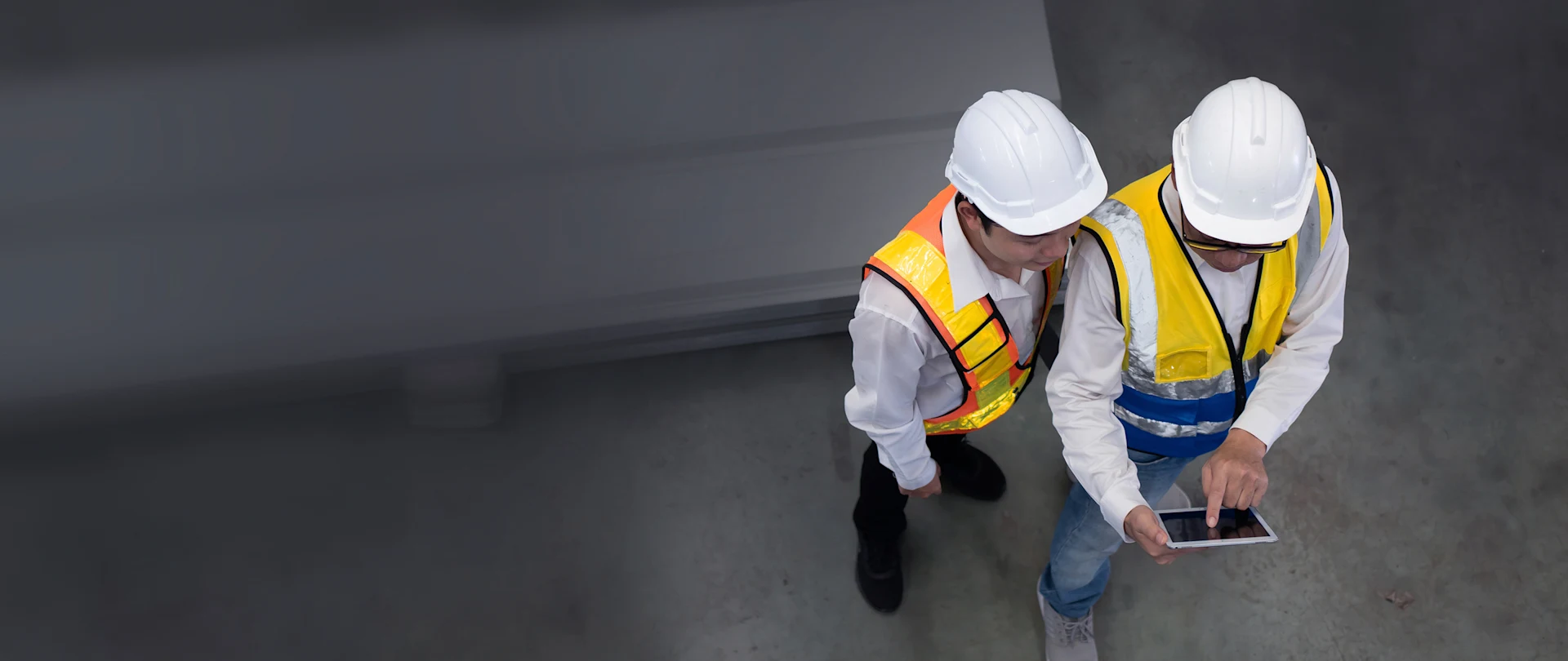 High-angle view of two workers in white hard hats and reflective safety vests using a tablet in an industrial facility.