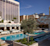 An upscale resort pool area in Omnia.jpg features lounge seating, cabanas, and umbrellas surrounding turquoise pools. A prominent green tree sits in a planter, with towering hotel buildings and a clear blue sky in the background.