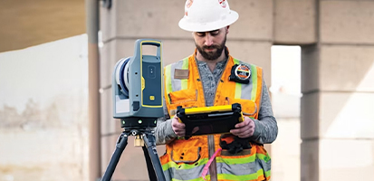 A construction worker in a hard hat and safety vest operates a Trimble X7 3D laser scanner with a rugged Trimble tablet on a job site.