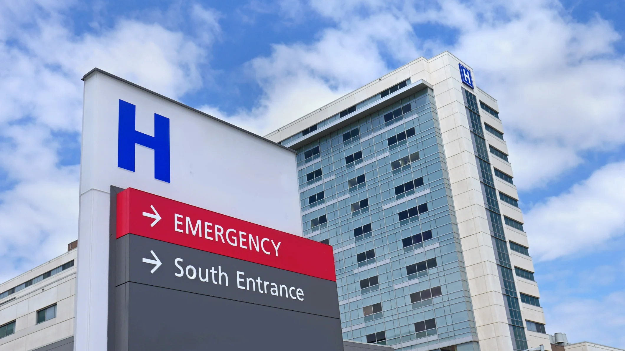 Modern hospital building, multi-story glass and light-colored panel facade. A prominent blue 'H' logo sign directs to 'EMERGENCY' and 'South Entrance'.