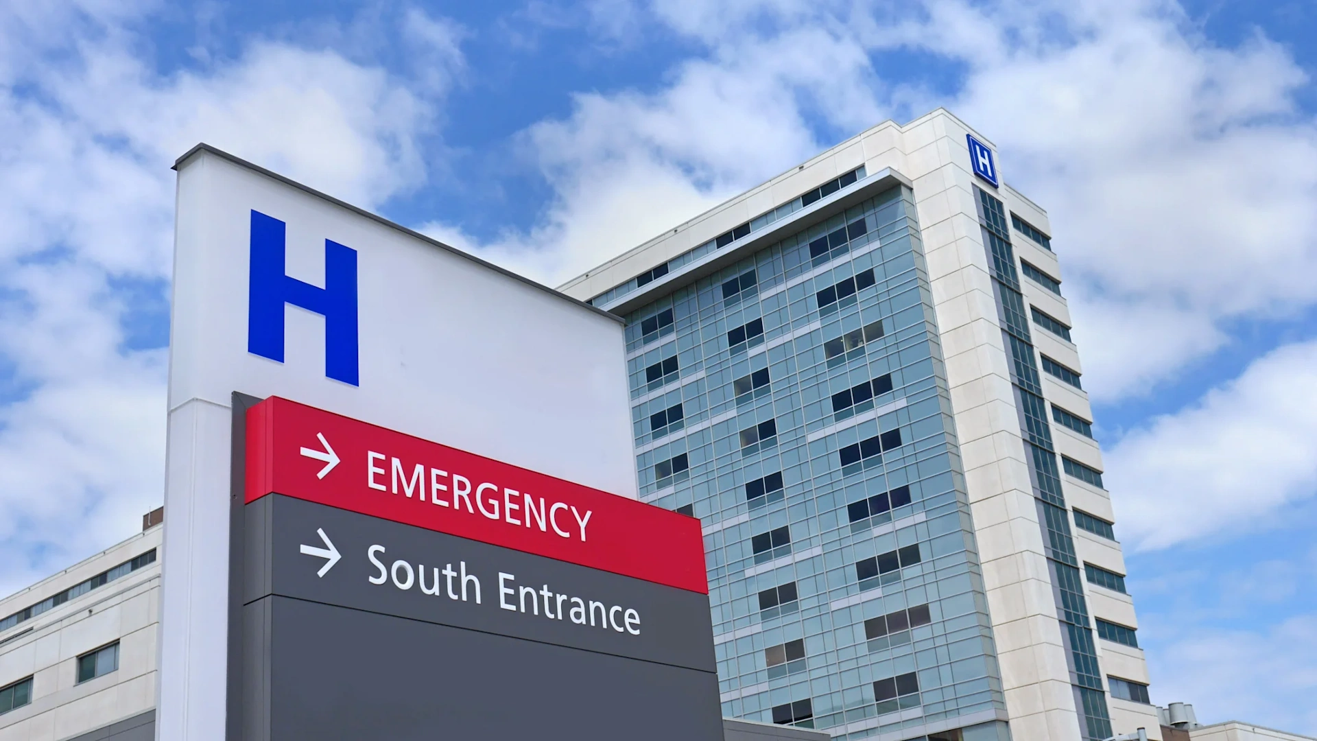 Modern hospital building, multi-story glass and light-colored panel facade. A prominent blue 'H' logo sign directs to 'EMERGENCY' and 'South Entrance'.